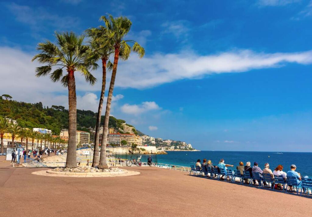 Passeggiata sulla Promenade des Anglais a Nizza con palme, sedie blu e vista sulla Baie des Anges